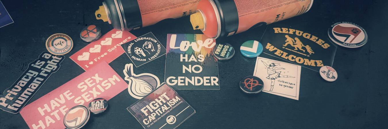 Banner format photo with a "retrolux" filter, making it look desaturated and "scratched". Depicted are two spraypaint cans, black and red, along with several LGBT+, relationship anarchy, veganism, clothes have no gender, anti-fascism and privacy buttons.